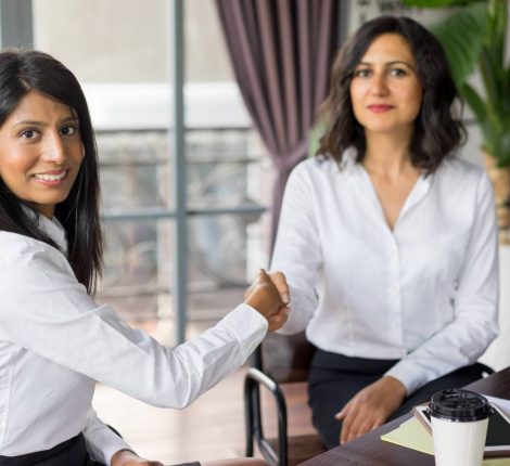 Portrait of successful female partners shaking hands. Young Latin American and Caucasian businesswomen sitting and greeting with handshake in office. Partnership concept