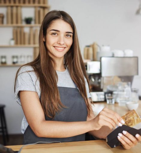 Female coffee shop employee using a credit card reader to bill the customer looking happy smiling at camera.